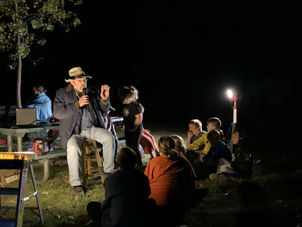 A man telling a story outside at night to a group of children gathered around his feet.