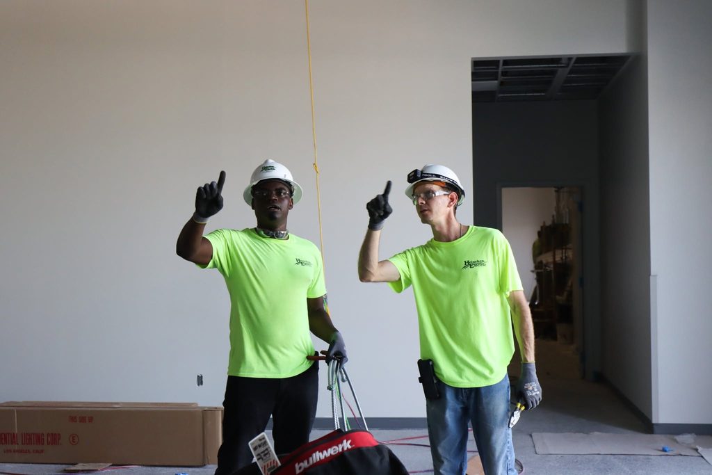 Two electric workers with hard hats point up as they discuss work.